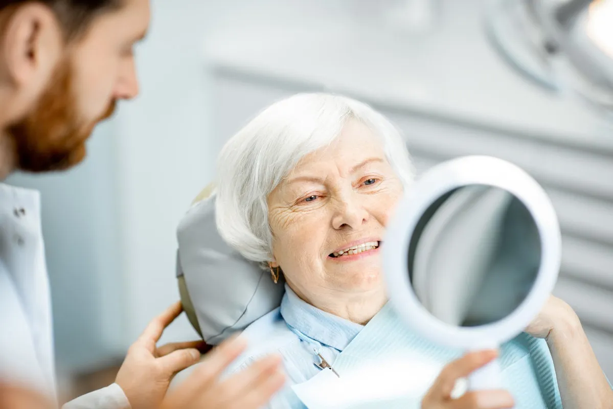 Happy elderly woman enjoying her beautiful toothy smile looking to the mirror in the dental office